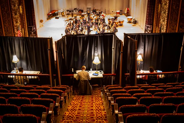 Adjudicators at lamplit desks in a theatre balcony overlooking a brass band on stage