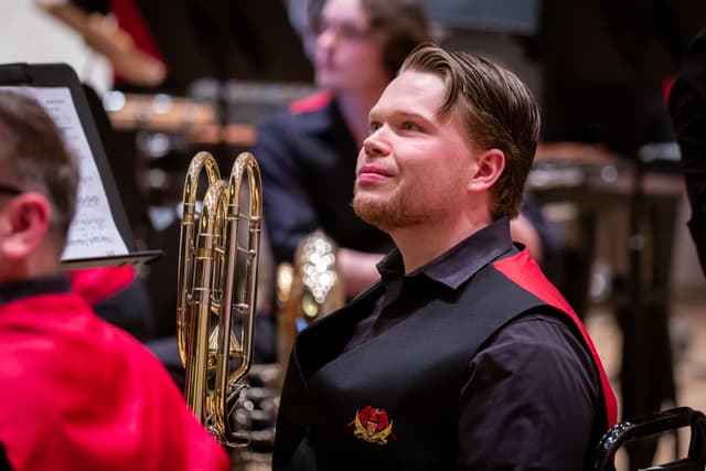 Seated brass band player in dark and red uniform looking up beside a trombone