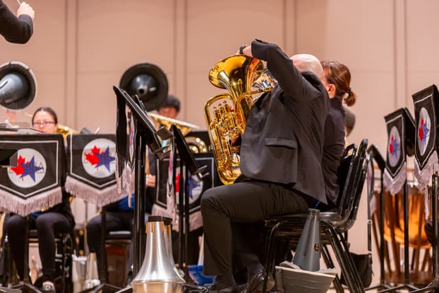 Euphonium player seated on stage among brass band music stands with banners, conductor's baton at top left.