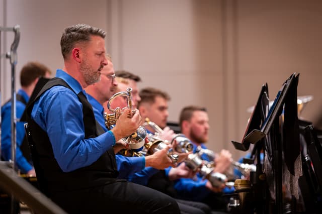 Brass band players in blue shirts seated in a row, a flugelhorn player in front