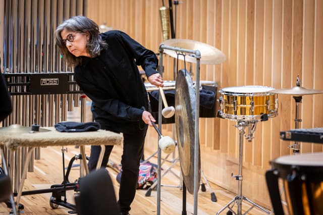 Percussionist holding mallets leans beside a gong, with chimes and snare drum in a wood-panelled room.