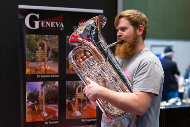 Musician playing a silver euphonium in front of a Geneva instruments banner at a trade stand