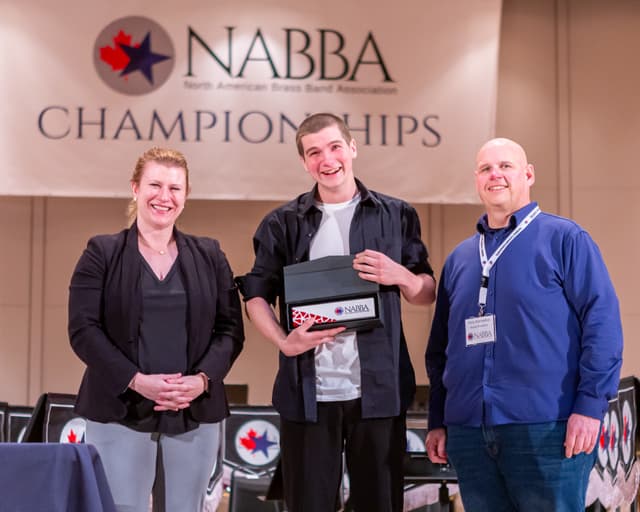 Three people smiling on stage with a boxed award beneath the NABBA Championships banner