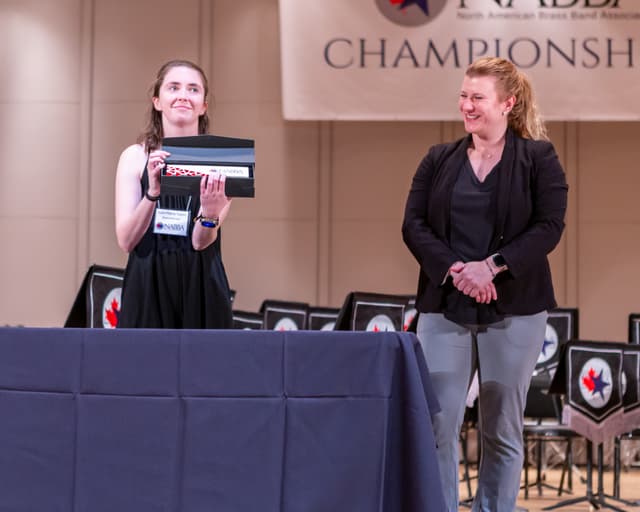 Two people on stage at the NABBA Championships presenting a boxed award beside a draped table