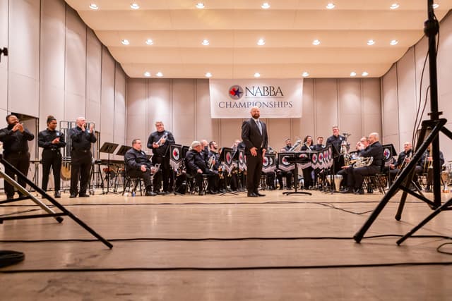 Conductor stands on stage as brass band members applaud beneath a NABBA Championships banner.