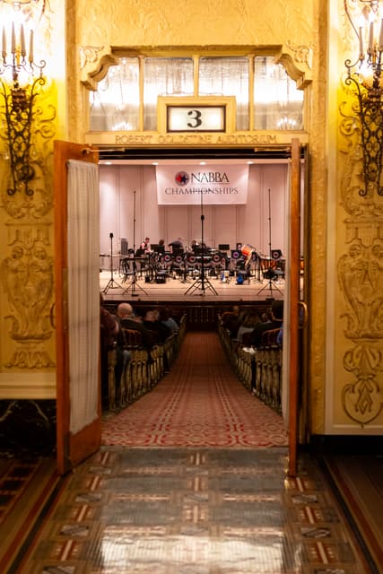 View through ornate doors into an auditorium with a NABBA Championships banner and a stage being set up