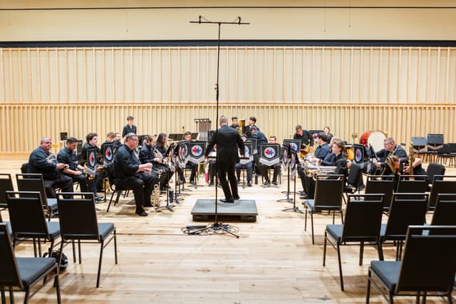Conductor on podium facing a seated brass band on a wood-panelled stage with empty chairs