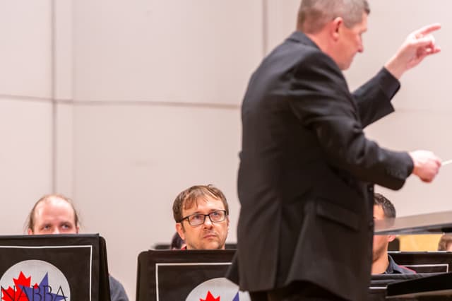 Conductor cueing the band while musicians watch from behind NABBA-branded music stands