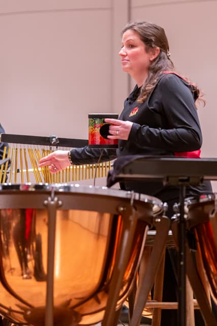 Percussionist holding a shaker beside timpani and bar chimes on a concert stage