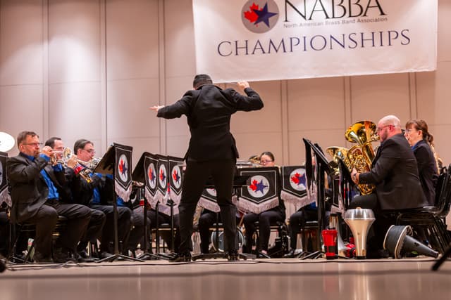 Conductor leading a brass band on stage beneath a NABBA Championships banner