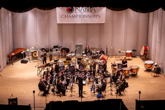 Brass band and conductor performing on stage beneath a NABBA Championships banner, percussion behind.