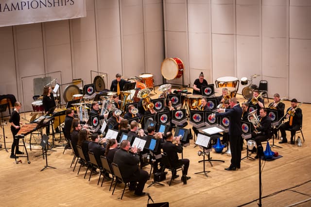 Brass band and conductor performing on stage with percussion behind, seen from above
