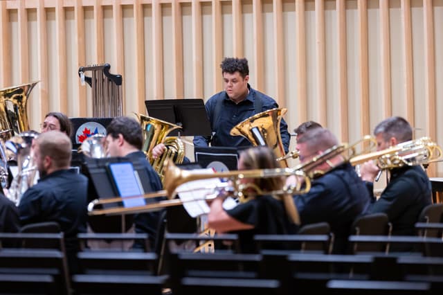 Brass band performing on stage with tubas and trombones, percussionist standing behind a music stand