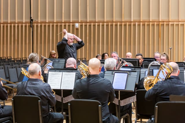 Conductor with raised arms leads a brass band in a wood-panelled concert hall