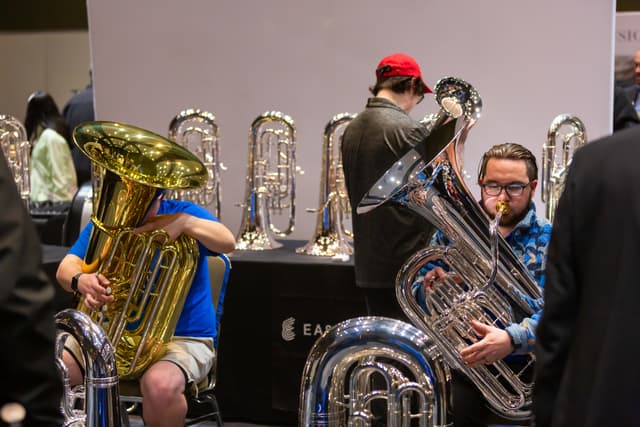 Two musicians test large tubas at an instrument trade display with silver instruments behind